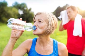 Senior woman drinking from water bottle outdoors with senior man wiping face with towel in background